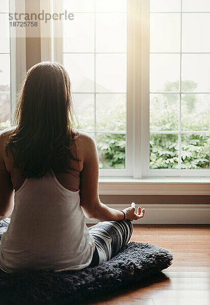 Frau sitzt drinnen auf dem Boden und meditiert vor Fenstern.