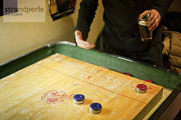 Ein Mann beugt sich über einen Vintage-Shuffleboard-Tisch aus Holz und hält ein Bier in einer Bar in Seattle  Washington.