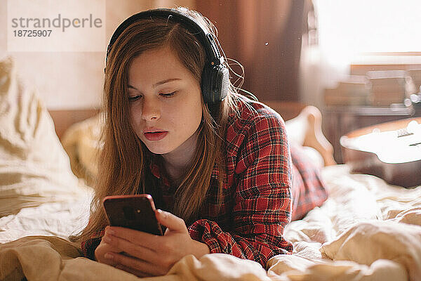 Teenager-Mädchen mit Kopfhörern und Smartphone zu Hause im Bett liegend