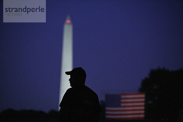 Silhouetten von Menschen  die an den leistungsstarken Lichtern vorbeigehen  die auf dem Washington Monument leuchten  und einer riesigen amerikanischen Flagge bei Nacht in Washington  D.C.