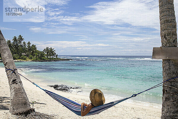 Mann mit Strohhut entspannt sich in der blauen Hängematte am Sandstrand von Samoa