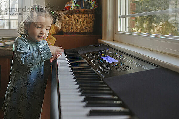 Ein süßes kleines Mädchen mit Zöpfen spielt auf einer Tastatur vor dem Fenster