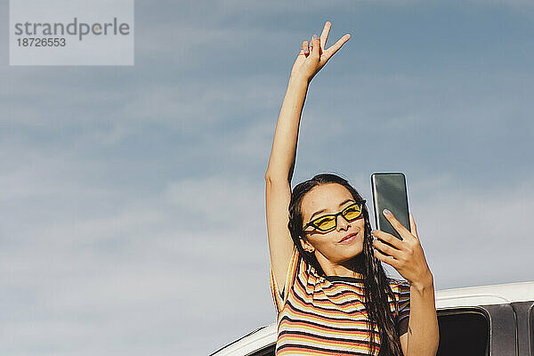 Junge Frau zeigt ein Friedenszeichen und macht ein Selfie per Smartphone