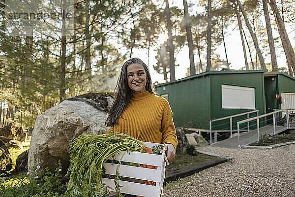 Reife Frau steht im Garten und trägt eine Kiste mit frischem Gemüse