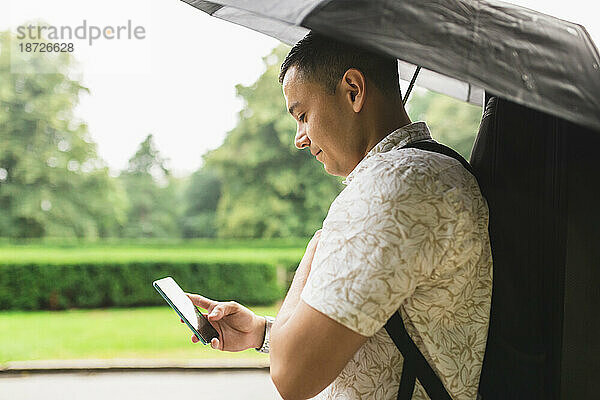 Junger Mann benutzt Smartphone und hält Regenschirm im Park