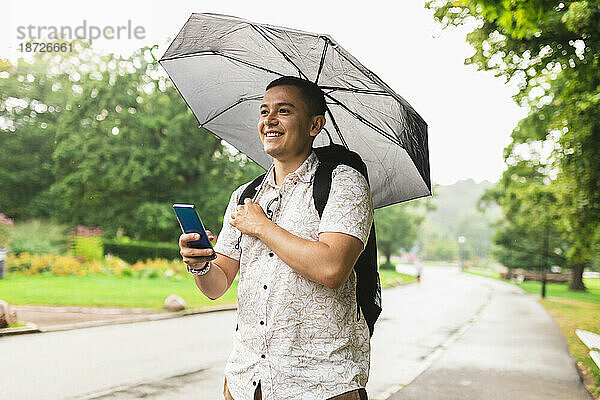 Junger Mann mit Regenschirm hält Smartphone im Park