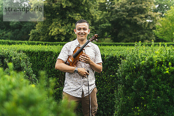 Lächelnder Mann mit Geige und Bogen steht vor Pflanzen im Park