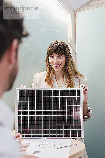 Lächelnde Geschäftsfrau zeigt einem Kollegen im Büro Solarpanel