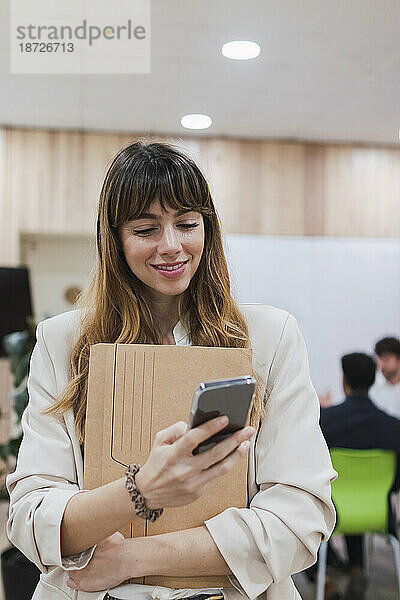 Lächelnde Geschäftsfrau hält Ordner in der Hand und benutzt Mobiltelefon im Büro
