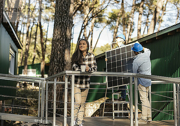 Photovoltaiktechniker montieren Solarpanel an nachhaltigem Haus  während der Hausbesitzer auf der Veranda steht