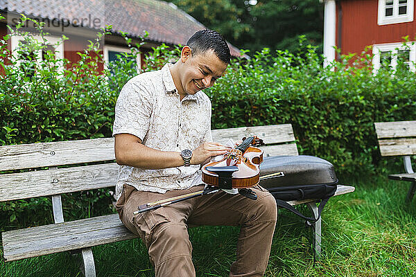 Smiling man cleaning violin sitting on bench at park