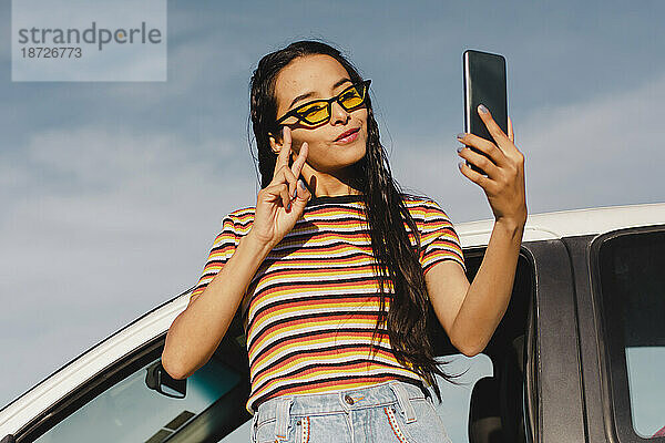 Lächelnde junge Frau macht ein Selfie mit ihrem Smartphone und lehnt sich ans Auto