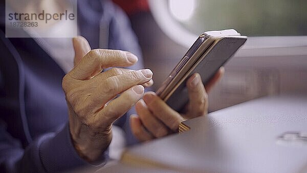 Nahaufnahme der Hände einer älteren Dame  die in einem Zugwaggon sitzt und ein Smartphone benutzt