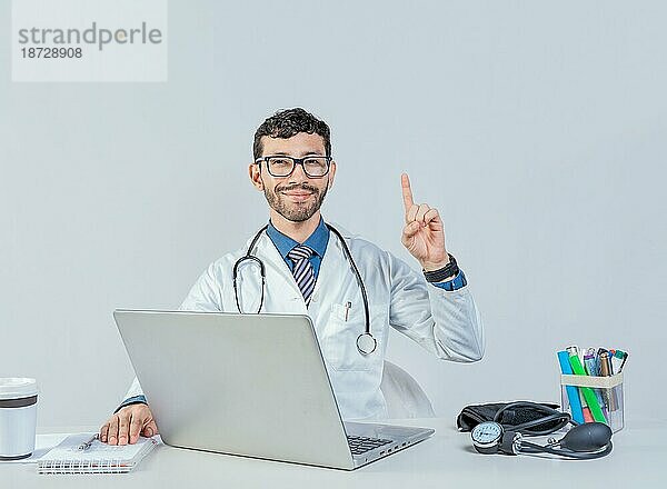 Lächelnder Arzt mit Laptop mit erhobenem Finger in der Idee Geste. Arzt sitzend mit Laptop nach oben zeigend isoliert. Junger Arzt sitzt mit Laptop mit einer Idee Geste