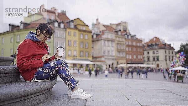 Ältere Dame sitzt auf den Stufen  trinkt Kaffee und benutzt ein Smartphone im historischen Zentrum einer alten europäischen Stadt. Schlossplatz  Altstadt von Warschau