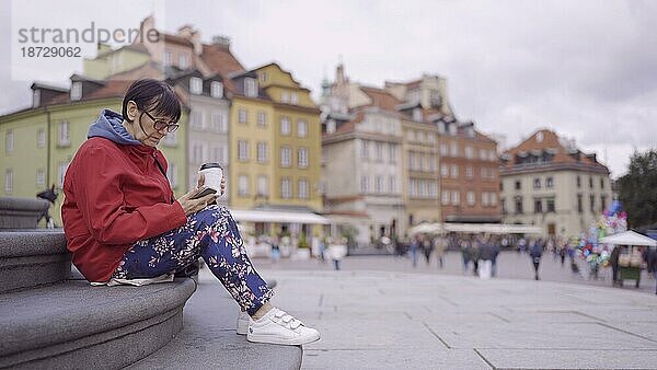 Ältere Frau sitzt auf den Stufen  trinkt Kaffee und benutzt ein Smartphone im historischen Zentrum einer alten europäischen Stadt. Schlossplatz  Altstadt von Warschau