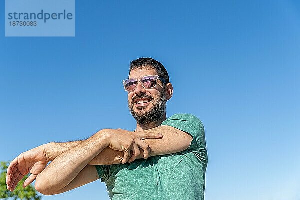 Vorderansicht eines gut aussehenden bärtigen Mannes mit Sonnenbrille  der lächelt und seine Muskeln auf einem Feld mit blauem Himmel im Hintergrund anspannt