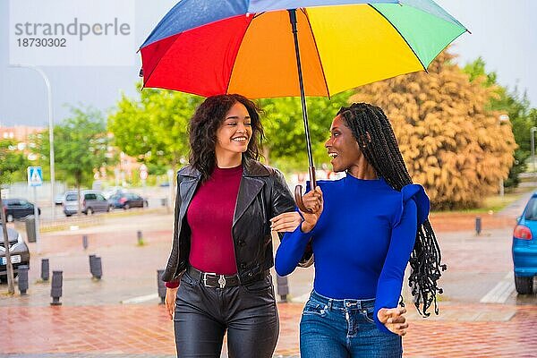 Multiethnische Freundinnen lachen mit einem Regenschirm im Regen in einer Stadt