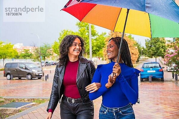 Multiethnische Freundinnen lachen mit einem Regenschirm im Regen in einer Stadt