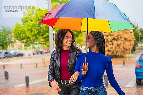 Porträt von lachenden multiethnischen Freundinnen mit einem Regenschirm im Regen in einer Stadt