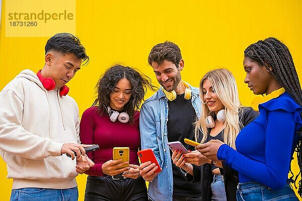 Niedriger Blickwinkel auf eine Gruppe lächelnder junger multiethnischer Teenager-Freunde  die Mobiltelefone auf einem gelben Hintergrund benutzen. Technologie süchtig Millennial Gemeinschaft Konzept. Social Media Kommunikation Generation Z