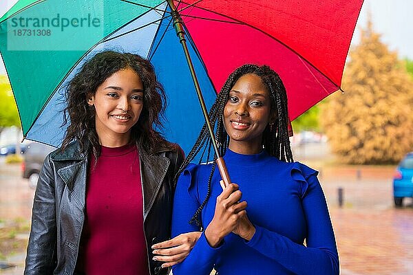 Porträt von lachenden multiethnischen Freundinnen mit einem Regenbogenschirm im Regen in einer Stadt