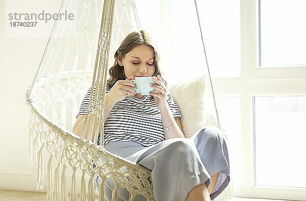 Schöne junge brünette Frau  die Tee oder Kaffee trinkt und eine Tasse mit beiden Händen hält  während sie sich in einem gestrickten Schaukelstuhl im Makramee Stil zu Hause am sonnigen frühen Morgen an einem faulen Wochenendtag entspannt