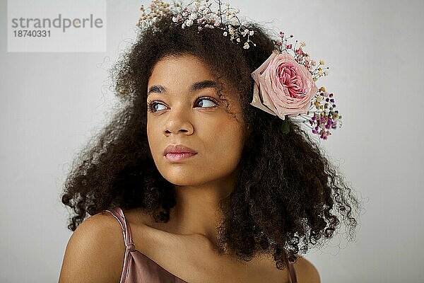 Close up Frühling Porträt der wunderschönen lächelnden afrikanisch amerikanischen jungen Frau mit frischen rosa Rose Blume in lockiges Haar  vorgrauem Hintergrund. Natürliche Schönheit der afrikanischen Frauen
