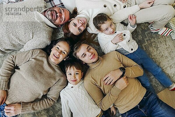 Draufsicht auf eine glückliche Mehrgenerationen Familie  die im Kreis auf dem Boden liegt und in die Kamera lächelt  um den Zusammenhalt zu stärken und von Kindheit an gesunde Beziehungen aufzubauen. Liebe und Zusammenhalt in der Familie