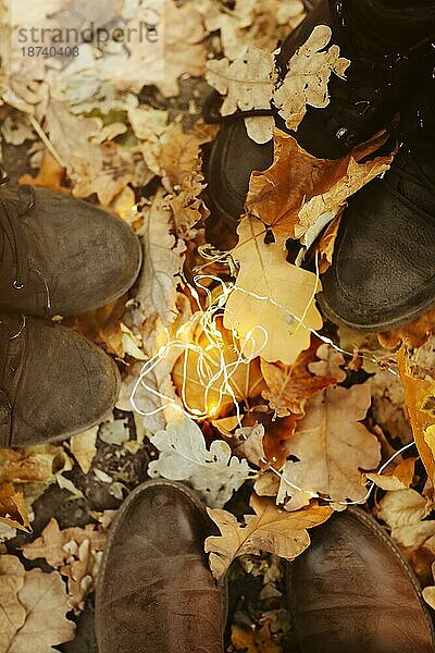 Von oben anonyme Menschen in Lederstiefeln  die auf gelben  verdorrten Blättern in der Nähe von leuchtenden Lichterketten in einem herbstlichen Park stehen