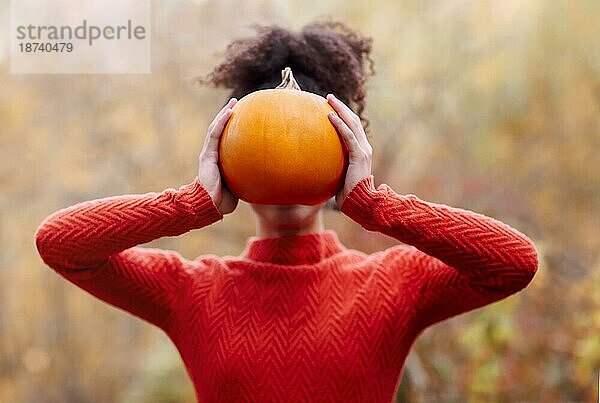 Junge gemischtrassige Frau mit lockigem Haar  die einen orangefarbenen Kürbis hält  während sie vor einem unscharfen Herbstwaldhintergrund steht. Herbstsaison und Ernte Konzept