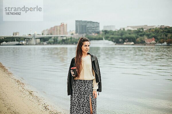 Junge Frau mit Kaffee zum Mitnehmen in der Hand in lässiger Kleidung am Flusswasser am Herbsttag mit Stadtlandschaft im Hintergrund  romantisch träumende Frau an der Küste  die Erholung in der Natur genießt