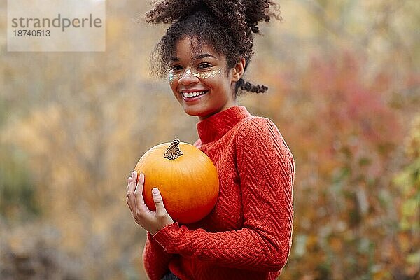 Junge glückliche fröhliche afroamerikanische Frau mit lockigem Haar hält orange Kürbis und lächelnd in die Kamera  während gegen unscharfen Herbst Wald Hintergrund stehen. Herbstsaison und Halloween Konzept