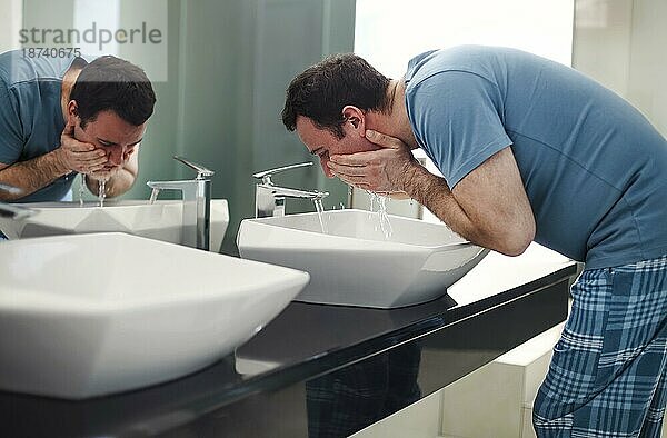 Brünetter Mann wäscht sein Gesicht in einem modernen Badezimmer am Morgen und trägt einen blaün Pyjama. Gesundheitswesen und Hygiene Konzept