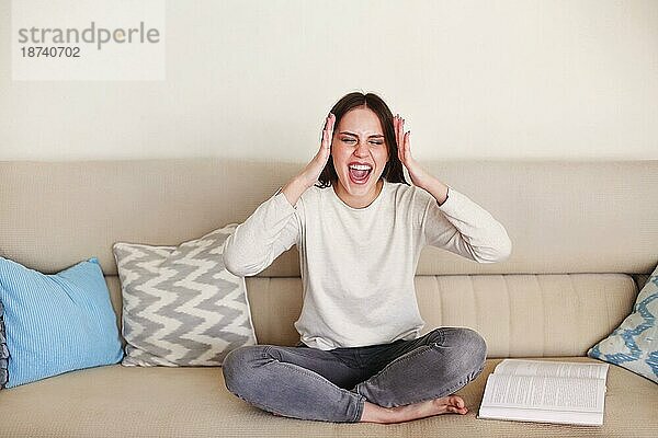 Genervte junge Frau  die auf dem Sofa sitzt und schreit  während sie zu Hause mit Laptop und Papieren arbeitet  während sie sich selbst isoliert