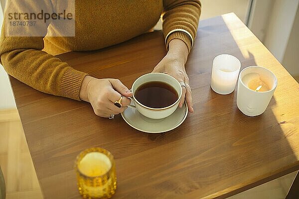 Ausgeschnittenes Foto einer Frau mit einer Tasse Tee  die an einem Tisch mit brennenden Kerzen sitzt  eine nicht erkennbare Frau hält eine Tasse mit einem heißen Morgengetränk  während sie an einem hölzernen Schreibtisch sitzt und sich zu Hause entspannt