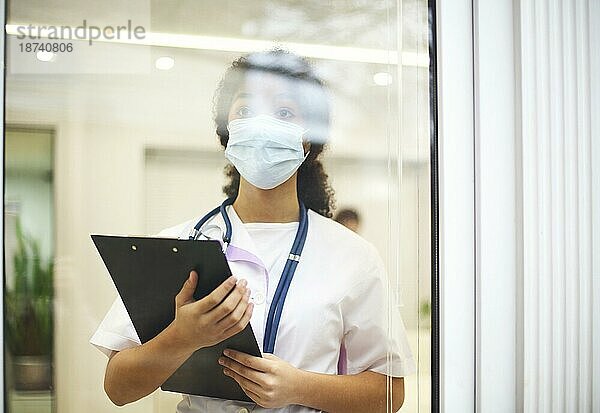 Junger müder  überarbeiteter afroamerikanischer medizinischer Angestellter mit Mundnasenschutz und Röntgenbild in der Hand  der mit frustriertem Gesichtsausdruck aus dem Fenster schaut. Mitarbeiter im Gesundheitswesen während der COVID 19 Pandemie
