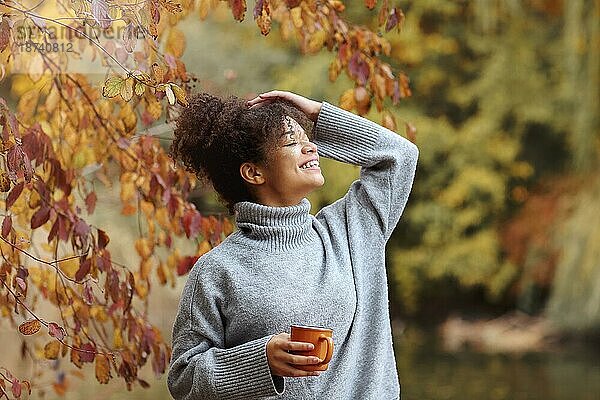 Junge fröhlich lächelnde gemischtrassige Frau mit Kaffeetasse in herbstlicher Natur  zufriedene Afroamerikanerin mit lockigem Haar im Strickpullover  die wegschaut  während sie den Morgen in einem schönen Herbstgarten genießt