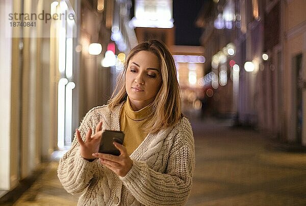 Junge attraktive Frau  die ihr Handy im Freien benutzt  eine Frau  die auf einer Straße in der Stadt spazieren geht und eine Nachricht auf einem modernen Smartphone tippt oder während eines Abendspaziergangs mit Freunden online chattet