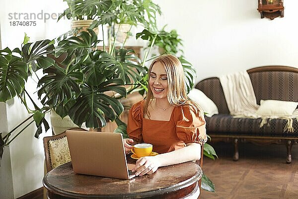 Fröhliche Frau  die an einem klassischen Tisch in einem hellen Wohnzimmer sitzt und mit der Hand am Laptop winkt  während sie ein Online Gespräch führt