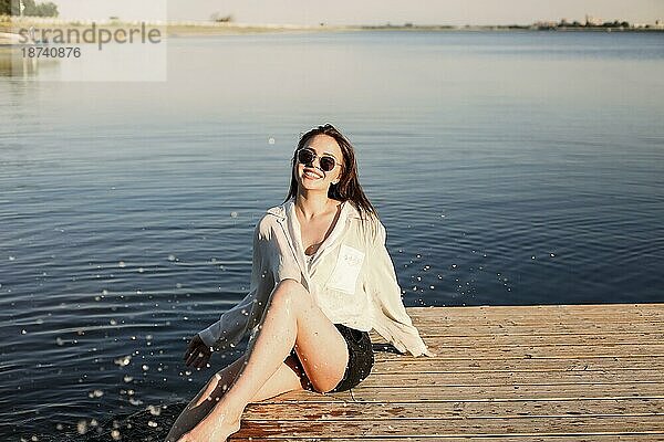 Seitenansicht einer jungen  unbekümmerten Frau mit modischer Kleidung und Sonnenbrille  die sich im Sommer auf einem Holzkai ausruht und in die Kamera schaut