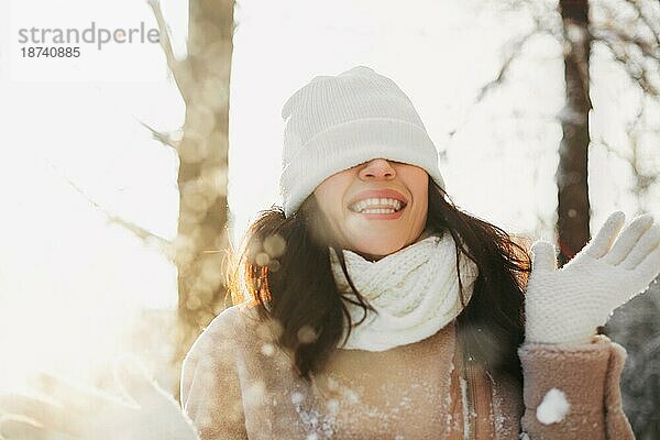 Optimistische junge Frau mit warmer Mütze auf den Augen lächelt  während sie auf der Straße an einem sonnigen Wintertag Spaß hat
