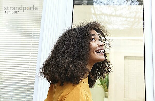 Fröhliche positive junge afroamerikanische Frau mit lockigem Haar  die aufschaut und lächelt  während sie in der Nähe des Fensters zu Hause steht  freudige fröhliche ethnische Millennial Frau  die das Gute und die Schönheit des Lebens schätzt