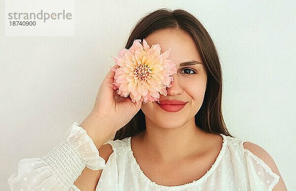 Fröhliche junge Frau schaut lächelnd in die Kamera und bedeckt ihr Auge mit einer natürlichen Blume vor weißem Hintergrund