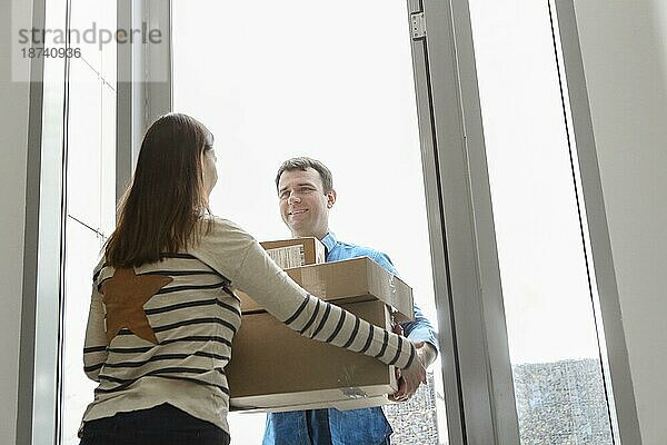 Schneller und zuverlässiger Service. Fröhlicher junger Kurier  der einer jungen Frau  die am Eingang zu ihrer Wohnung steht  einen Karton übergibt