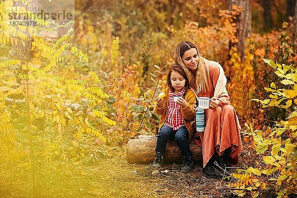 Junge Familie  Mutter und kleine Tochter  in stilvoller Kleidung genießen ein heißes Getränk  während sie im Herbst Wald sitzen