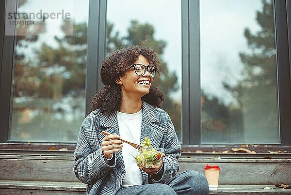 Glückliche afroamerikanische Frau Büroangestellte ißt Salat und lächelt in die Kamera  während sie auf einer Bank im Park im Freien sitzt  selektiver Fokus. Positives schwarzes Mädchen beim Mittagessen draußen während der Pause bei der Arbeit