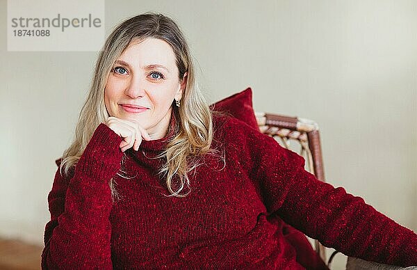 Attraktive glückliche Frau mittleren Alters  die auf einem bequemen Stuhl zu Hause am Wochenende sitzt und sich entspannt  trägt einen bunten gestrickten warmen roten Pullover und lächelt in die Kamera. Menschen Lebensstil Lebensstil