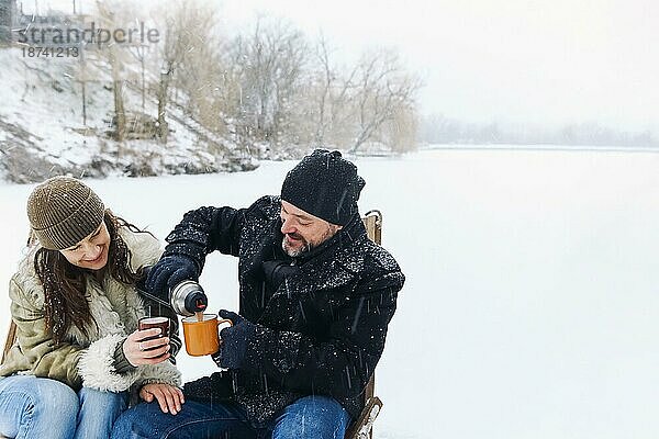 Glückliches  hübsches Paar mittleren Alters in warmer Kleidung  das während eines romantischen Wintertreffens in der Natur heißen Tee trinkt  ein verliebter Mann und eine verliebte Frau  die auf einem zugefrorenen See unter fallendem Schnee sitzen und sich an einem kalten  frostigen Tag unterhalten