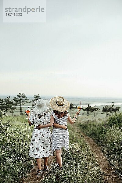Rückenansicht von anonymen Damen mit Alkoholcocktails die sich gegenseitig umarmen und auf einem Pfad an einem Sommertag in der Natur spazieren gehen